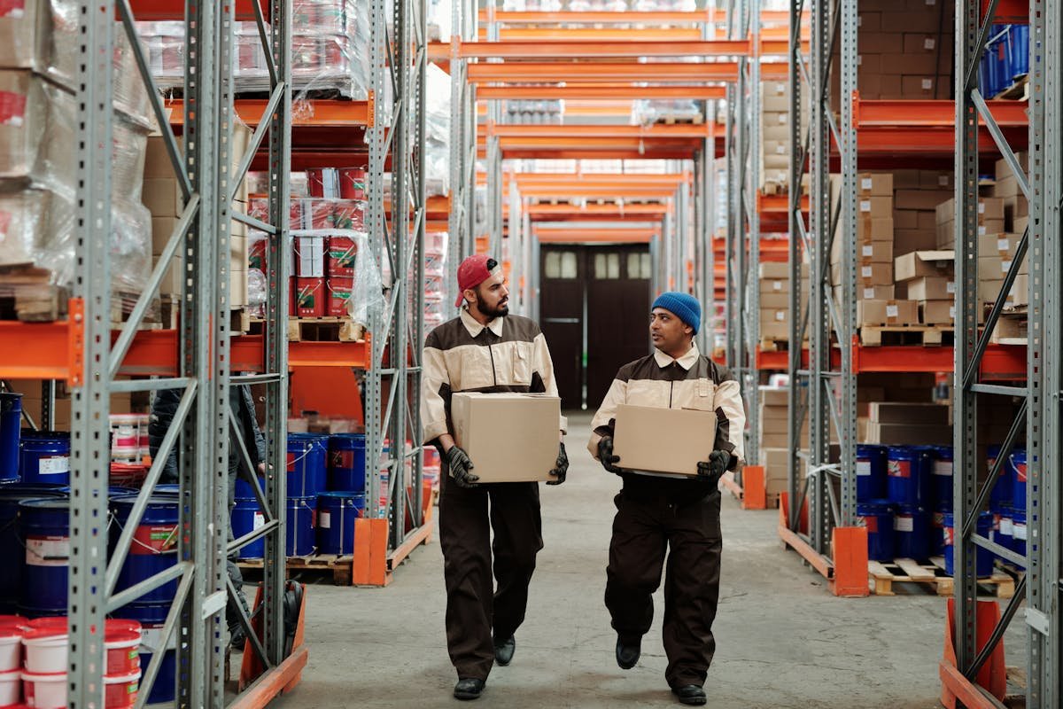 Two warehouse workers carrying boxes through a distribution centre aisle